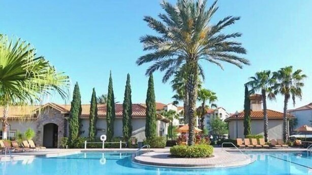 Piscina com palmeiras e espreguiçadeiras, cercada por edifícios com telhados vermelhos.