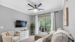 Quarto com televisão, ventoinha no teto, janela com cortinas, planta e cama com almofadas.