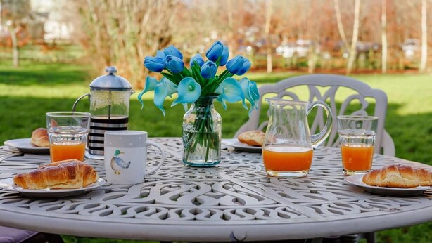 A round table with a glass pitcher, a glass with orange juice, a glass with tea, a mug with a bird design, and croissants on plates. - 2 Bed in Wrexham (oc-43916) (Overton)