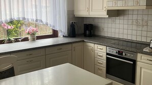 Une cuisine avec des meubles blancs, un plan de travail noir, un four encastré et une fenêtre ornée de rideaux légers.