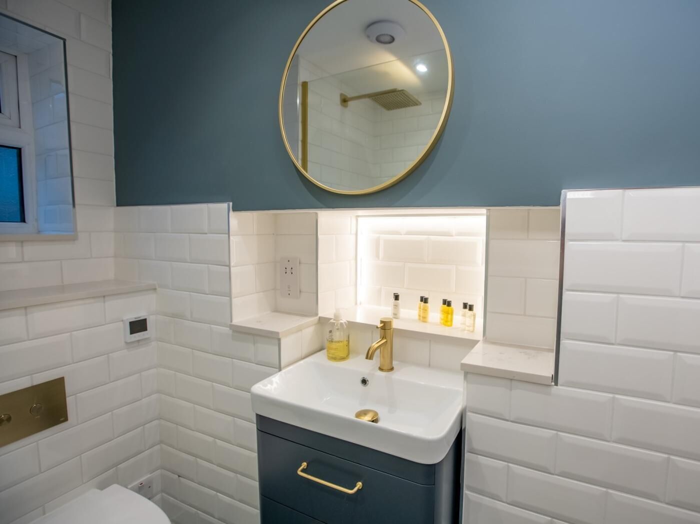 Et badeværelse med hvide metrofliser, et spejl med gylden ramme, en hvid vask med gylden vandhane og en hylde med toiletartikler.