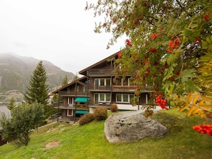 A two-story wooden chalet with a balcony, surrounded by greenery and a mountain backdrop. - Silence / Studio Türkis, (Grächen), 1024 by Interhome (Grächen)