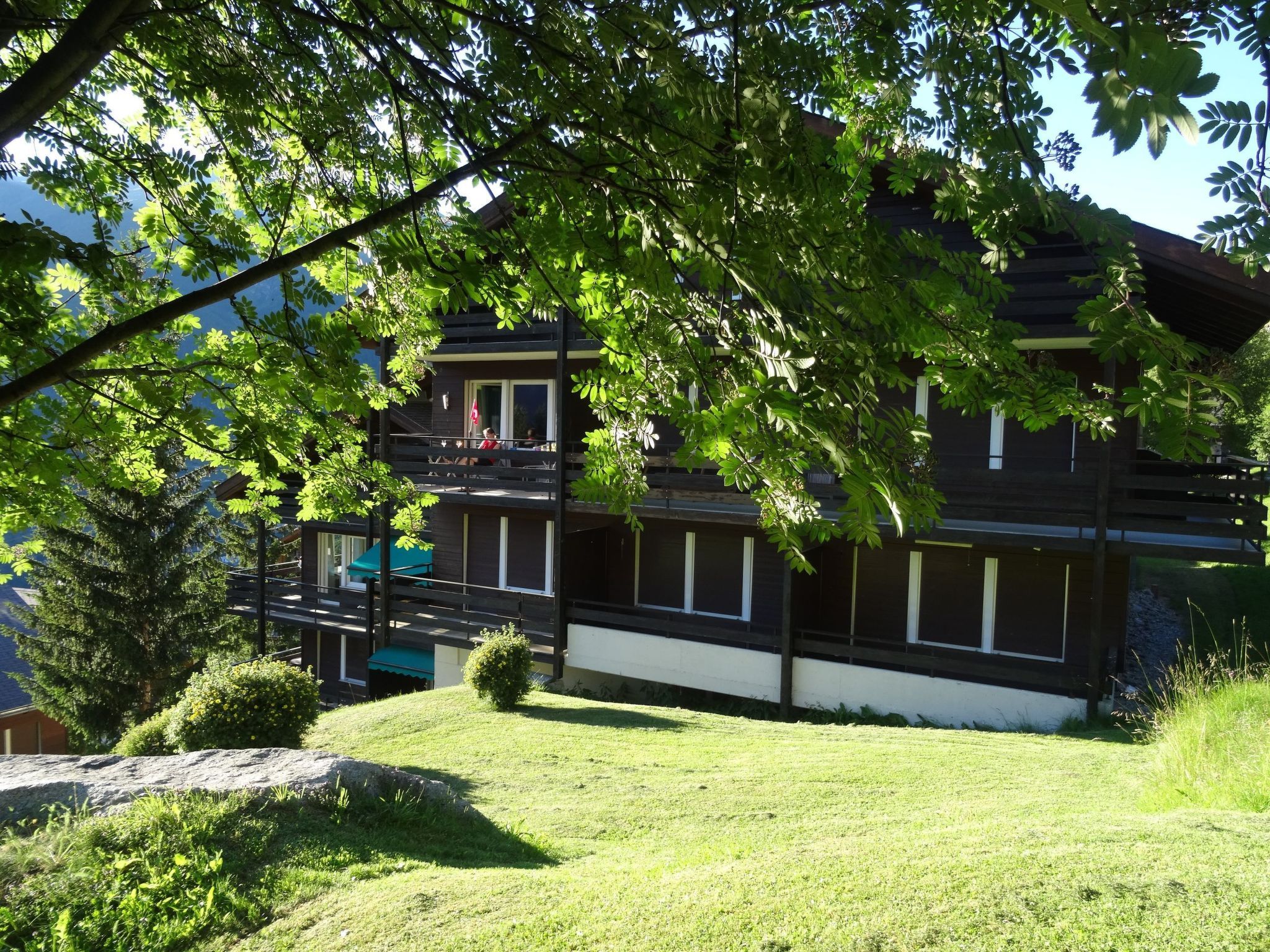 A two-story building with a balcony, surrounded by greenery and a grassy area.