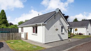 A small, single-story house with a sloped roof, a fenced yard, and a paved driveway. - 2 Bed in Inverness (oc-44481) (Drumnadrochit)
