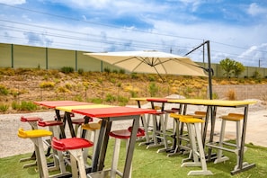 Mobiliario exterior con mesas y sillas, un paraguas grande y una zona ajardinada con un muro al fondo.