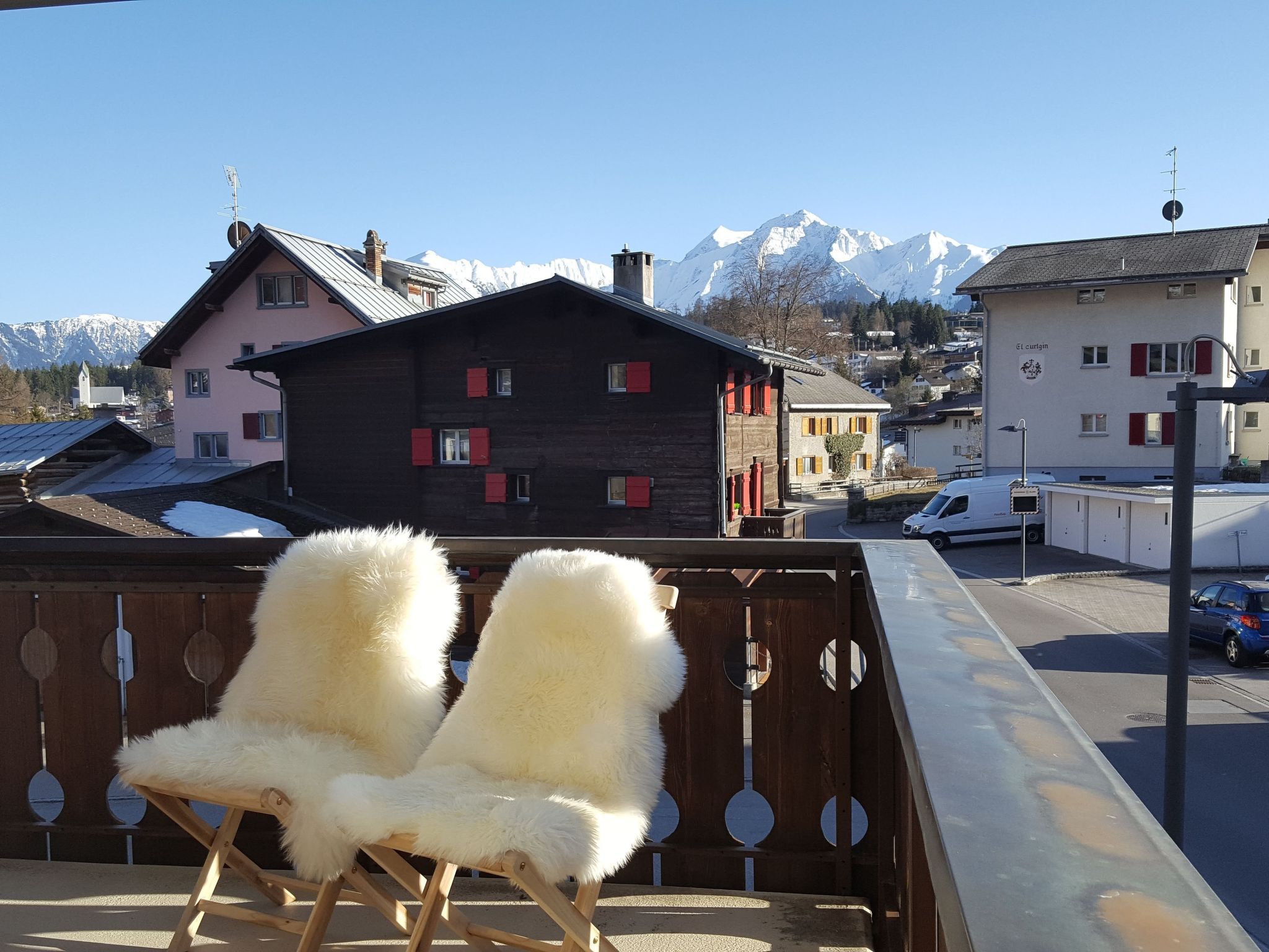 A balcony with two white furry chairs, a view of a mountain, and a building with red shutters.