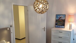 Makuuhuone, jossa on sänky, valkoinen lipasto laatikoineen, riippuvalaisin, ovi ja lipaston päällä oleva kehystetty kuva.