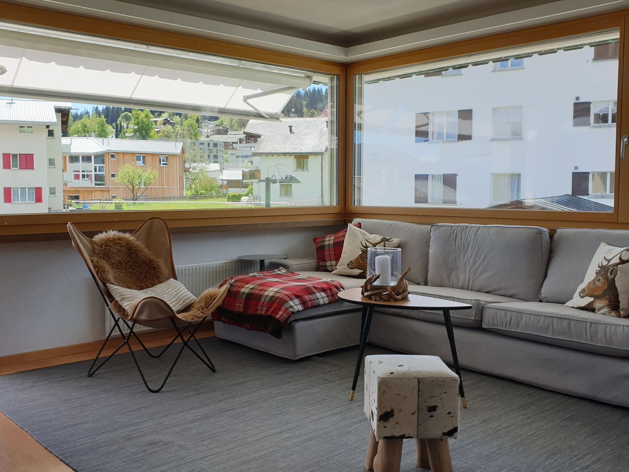 A modern living room with a sofa, a chair, a small table, and a view of buildings through large windows.