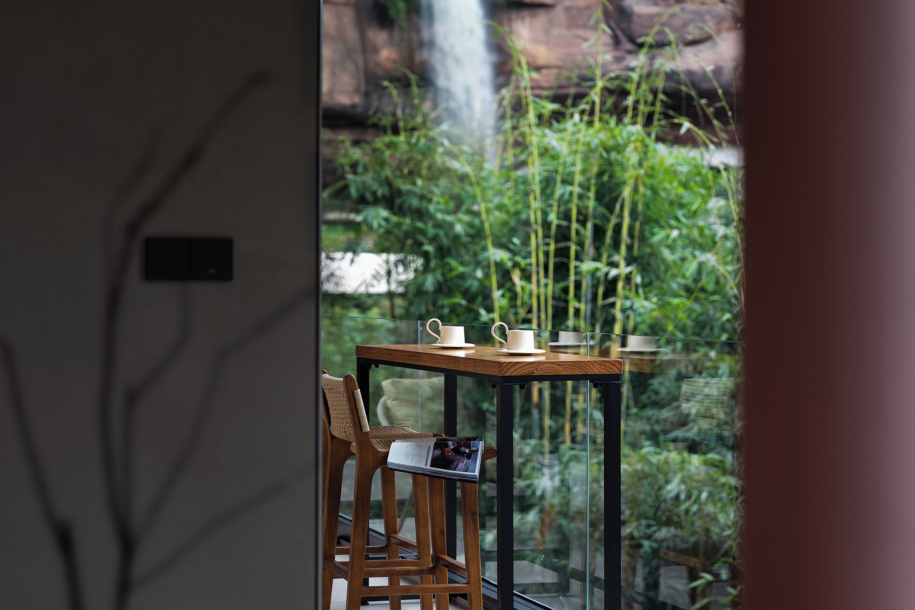 A table with cups and saucers, a chair, and a view of greenery and a waterfall.