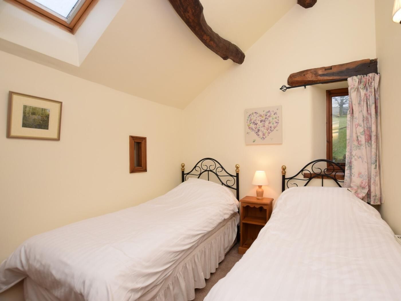 A bedroom with two single beds, a small bedside table with a lamp, a framed picture on the wall, and a window with curtains.