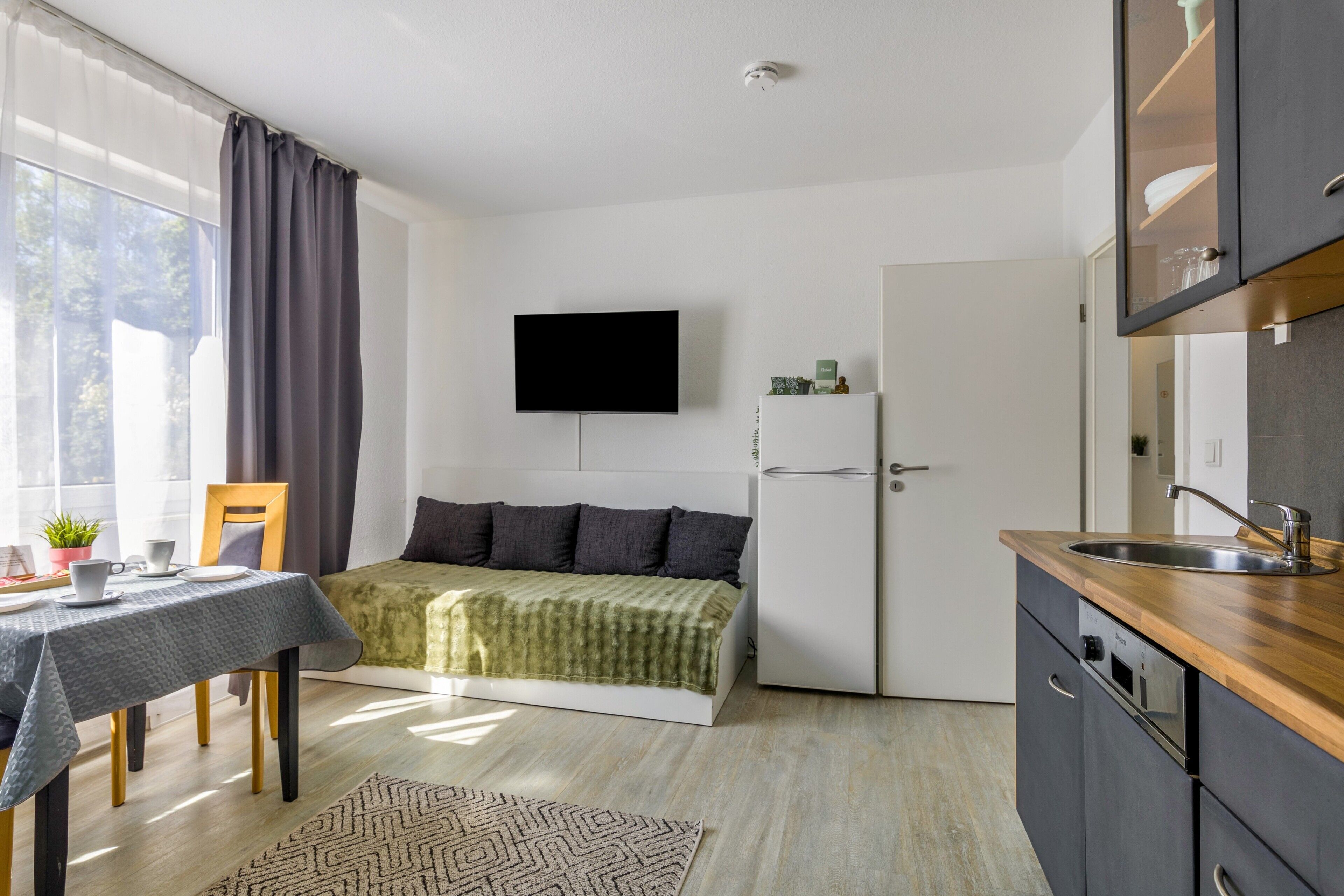 Une pièce de taille modeste comprenant un lit, une petite table avec des tasses, une chaise, un téléviseur fixé au mur, un réfrigérateur et un coin cuisine avec un évier et des placards.