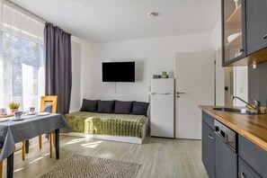 A compact room with a bed, a small table with cups, a chair, a TV mounted on the wall, a refrigerator, and a kitchen area with a sink and cabinets.