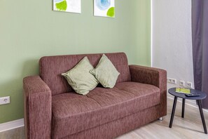 A room with a brown sofa, two pillows, a small black table with a book and remote, and a green wall with two abstract paintings. - Apartment 'Rhein Living Business' with Wi-Fi (Duisburg)