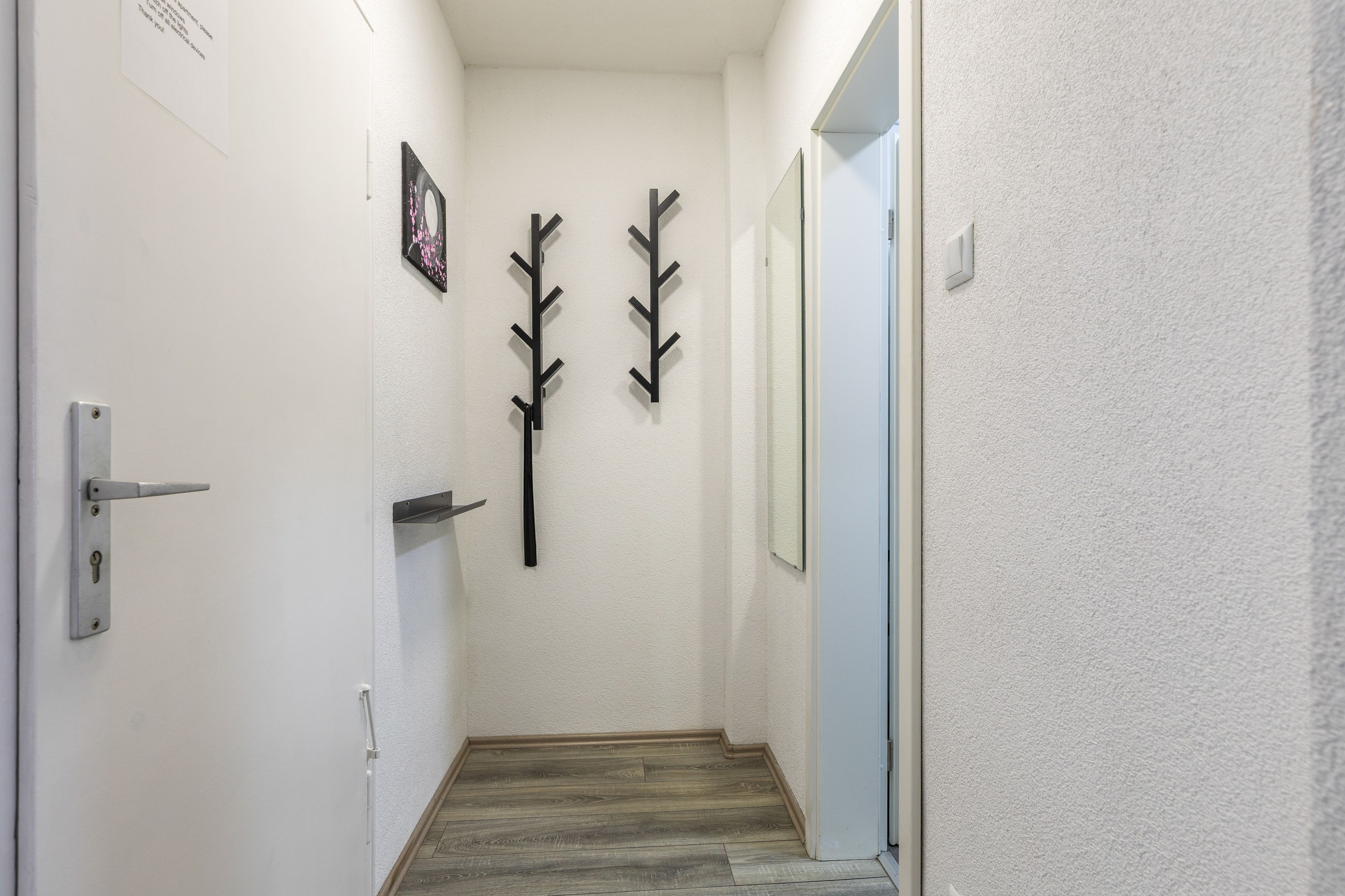 A narrow hallway with a wooden floor, white walls, and a door on the left. There are two black wall-mounted hooks on the wall.