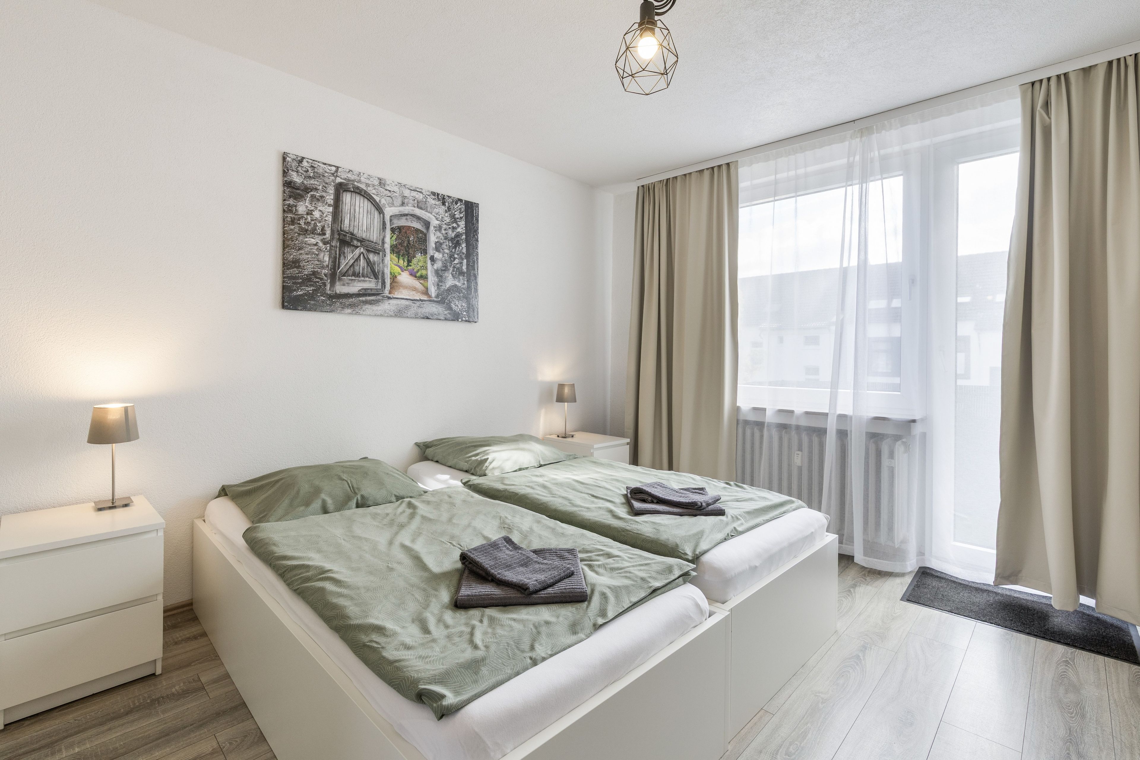 Habitació d'hotel amb dos llits, un quadre a la paret, una finestra amb cortines i una làmpada a cada costat.