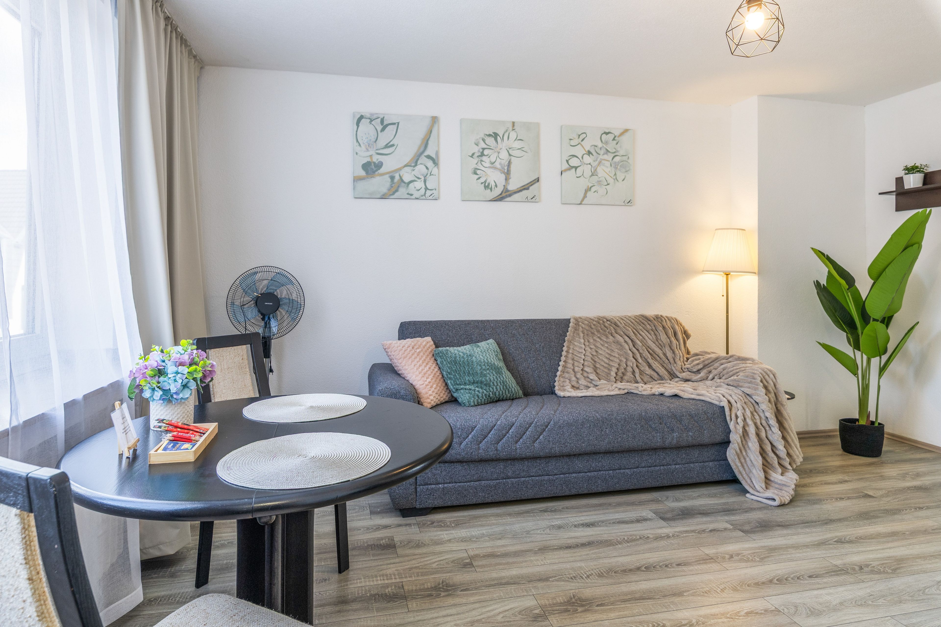 A living room with a grey sofa, a round dining table with placemats, a potted plant, and wall art.