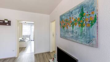 A modern living room with a flat-screen TV, a painting of flowers, and a hallway leading to a dining area.