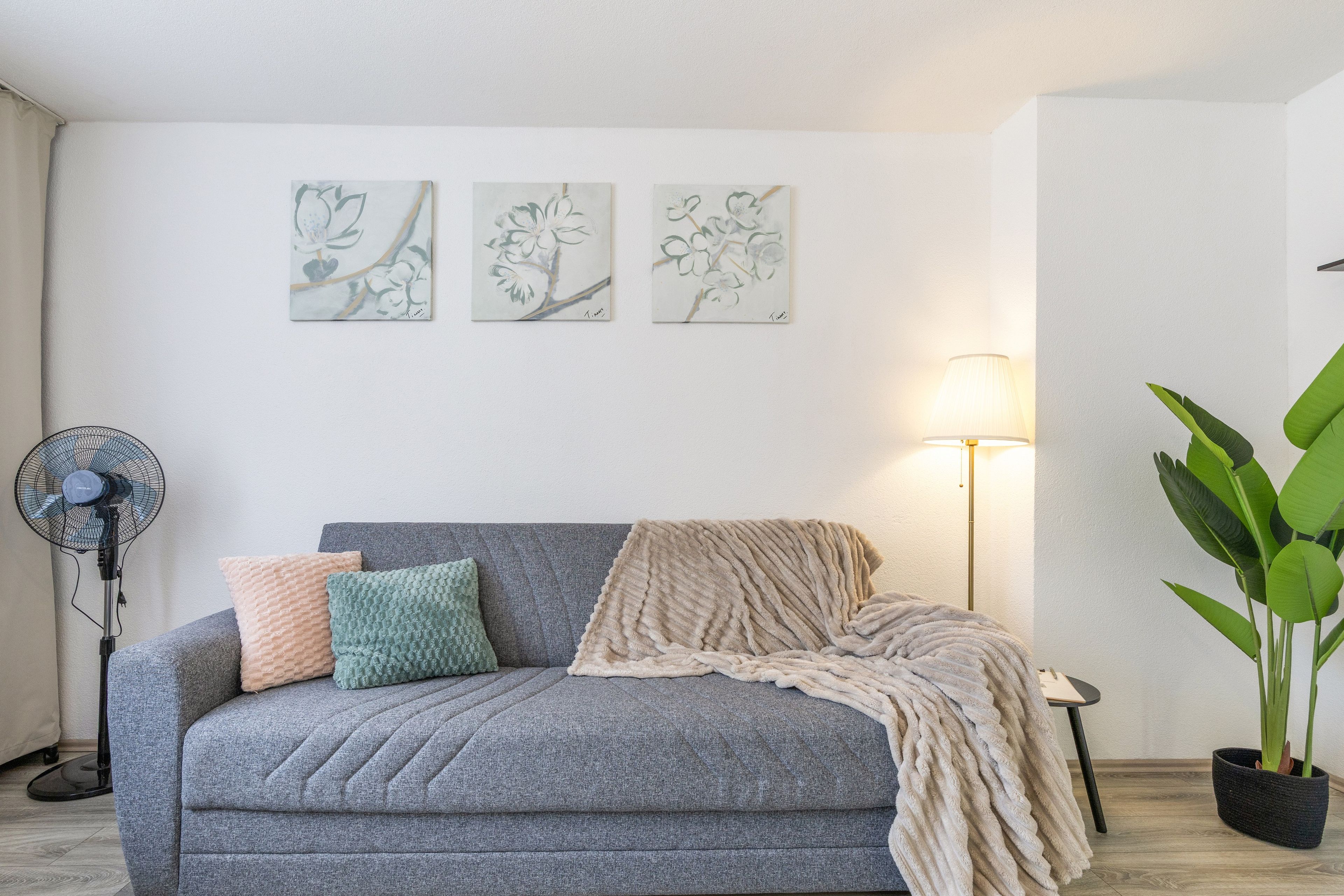 Una sala de estar con un sofá gris, un ventilador, una lámpara y una planta en maceta.