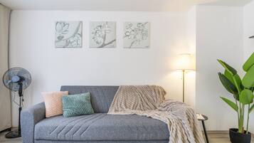 Una sala de estar con un sofá gris, un ventilador, una lámpara y una planta en maceta.