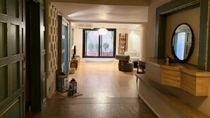 Eine Hotelhalle mit einem Holzbuchttisch, einem großen Spiegel, einer Sitzecke mit einer Couch und Sesseln, einem kleinen Tisch mit einer Lampe und Blick ins Freie durch eine Glastür.