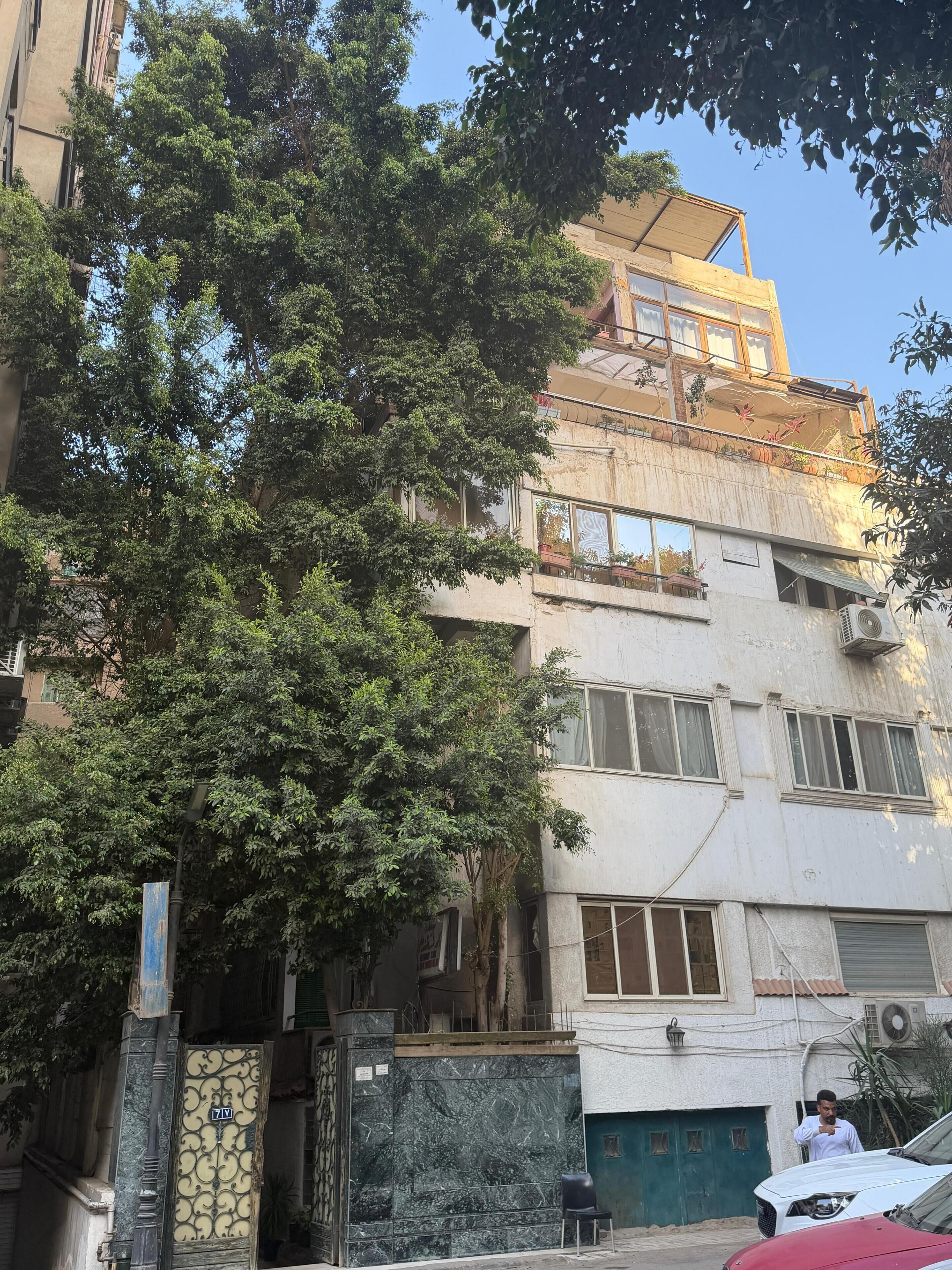 Un immeuble à plusieurs étages doté d’un balcon, avec une voiture garée devant et un arbre au premier plan.
