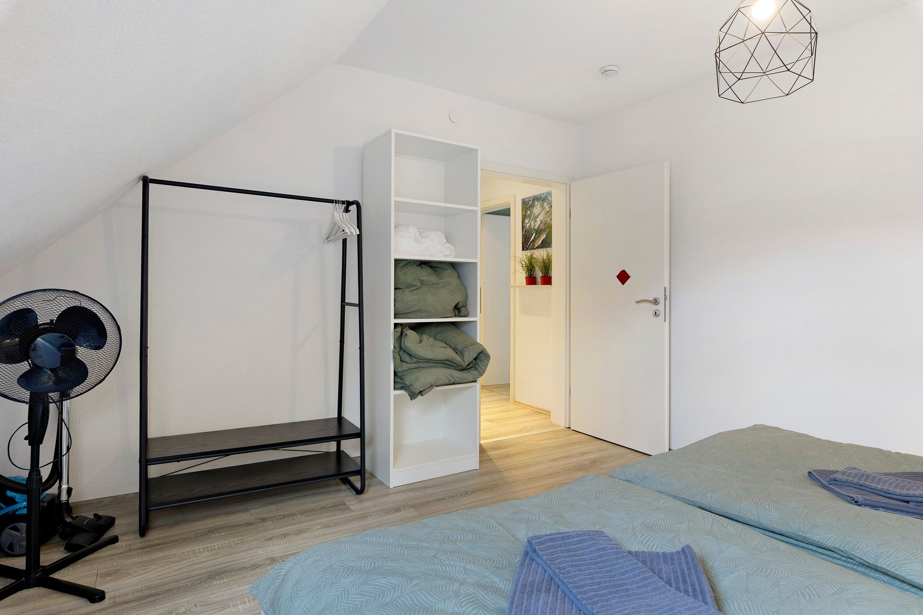 A bedroom with a bed, a standing fan, a vacuum cleaner, and a wardrobe.