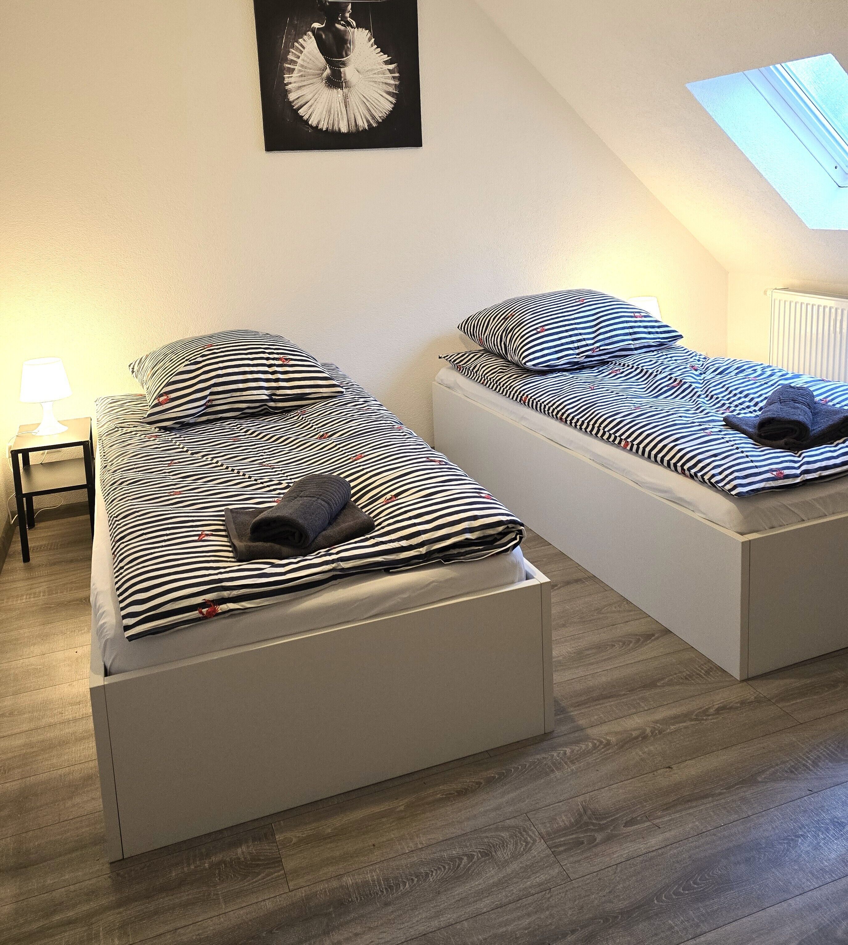Duas camas de solteiro com roupa de cama listrada, uma mesa de cabeceira com abajur e uma obra de arte emoldurada na parede.