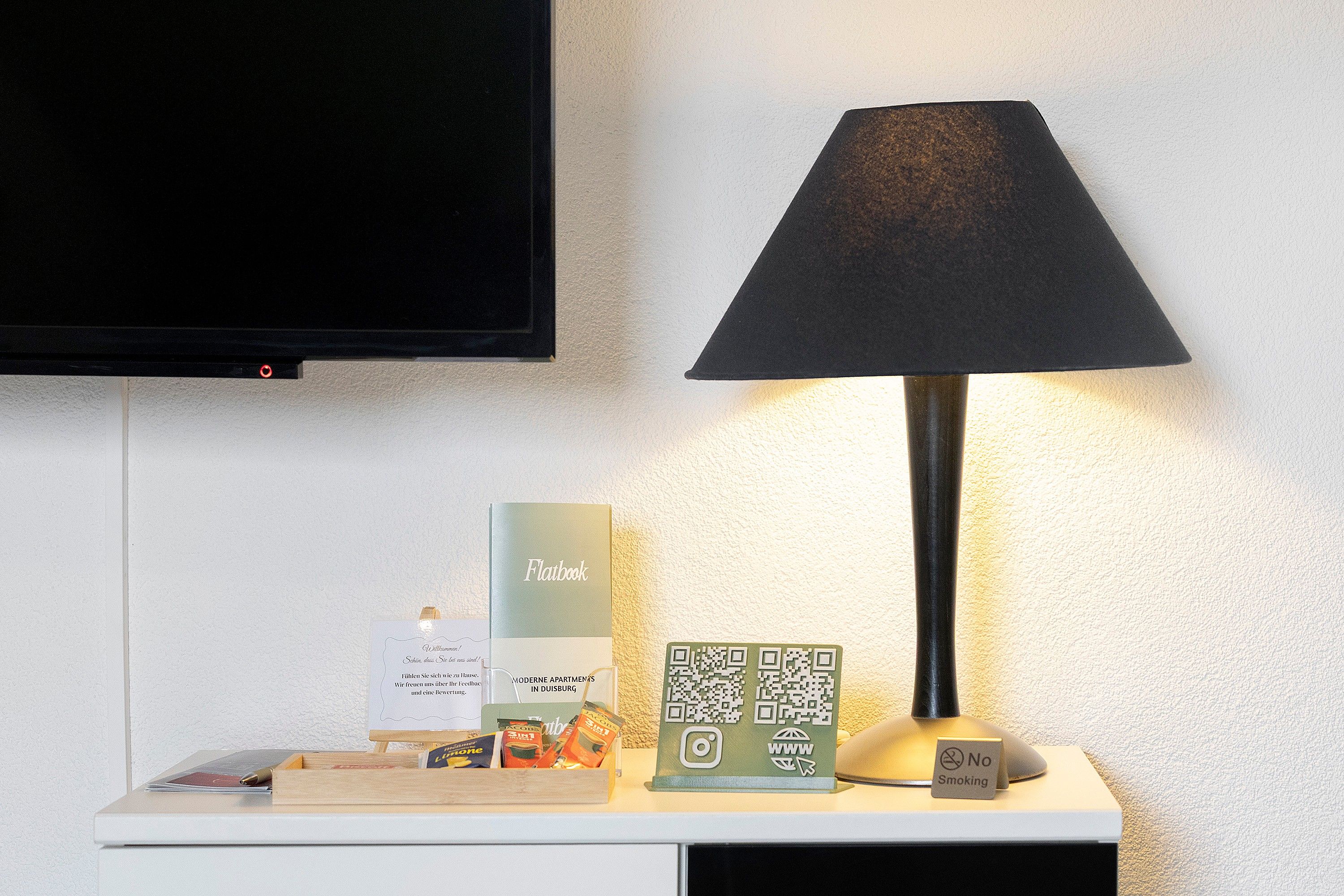 Un televisor de pantalla plana montado en la pared, una lámpara de mesa negra, un libro plano, un código QR y un letrero de 'No Fumar'.