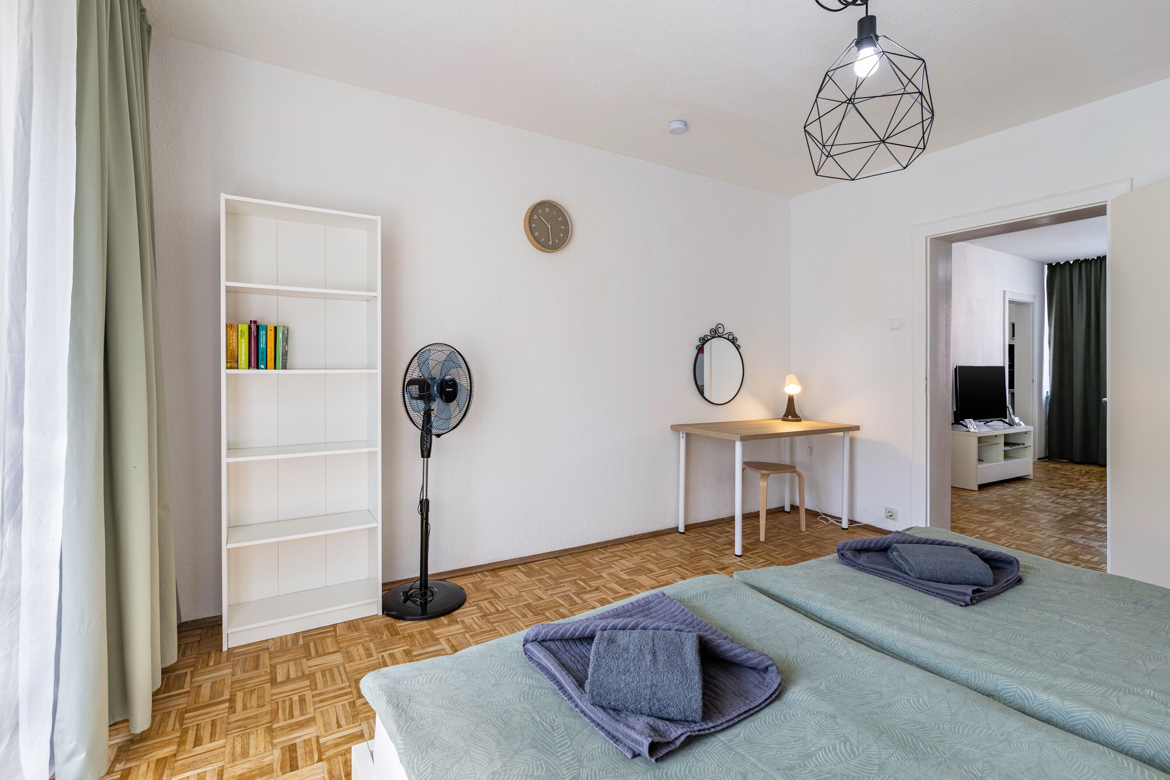 Hotellihuone, jossa on sänky, lattiatuuletin, työpöytä lampun kanssa, kirjahylly ja peili.
