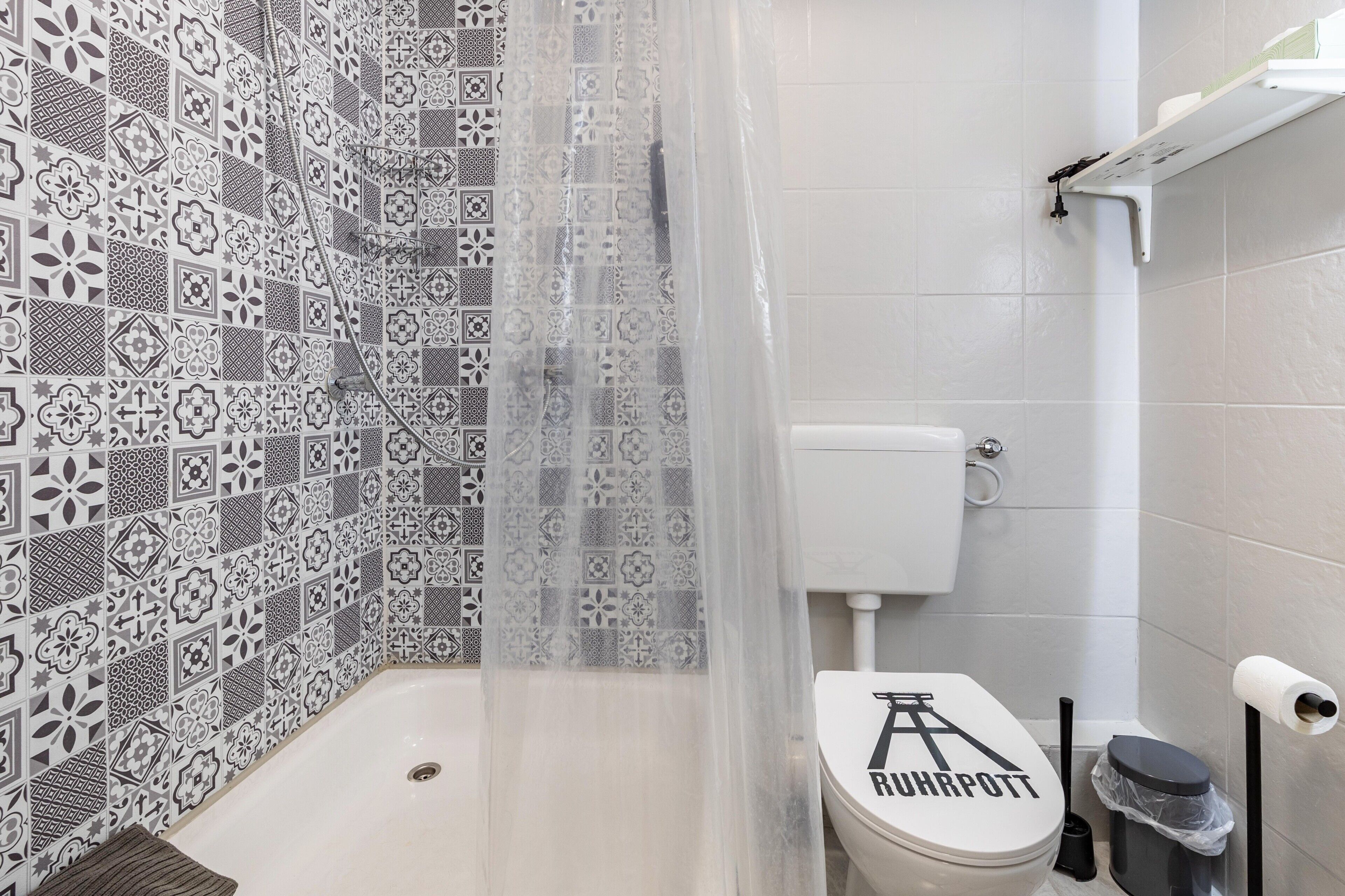 A bathroom with patterned tiles, a toilet with a 'RUHRPOTT' label, a shower curtain, and a toilet brush.