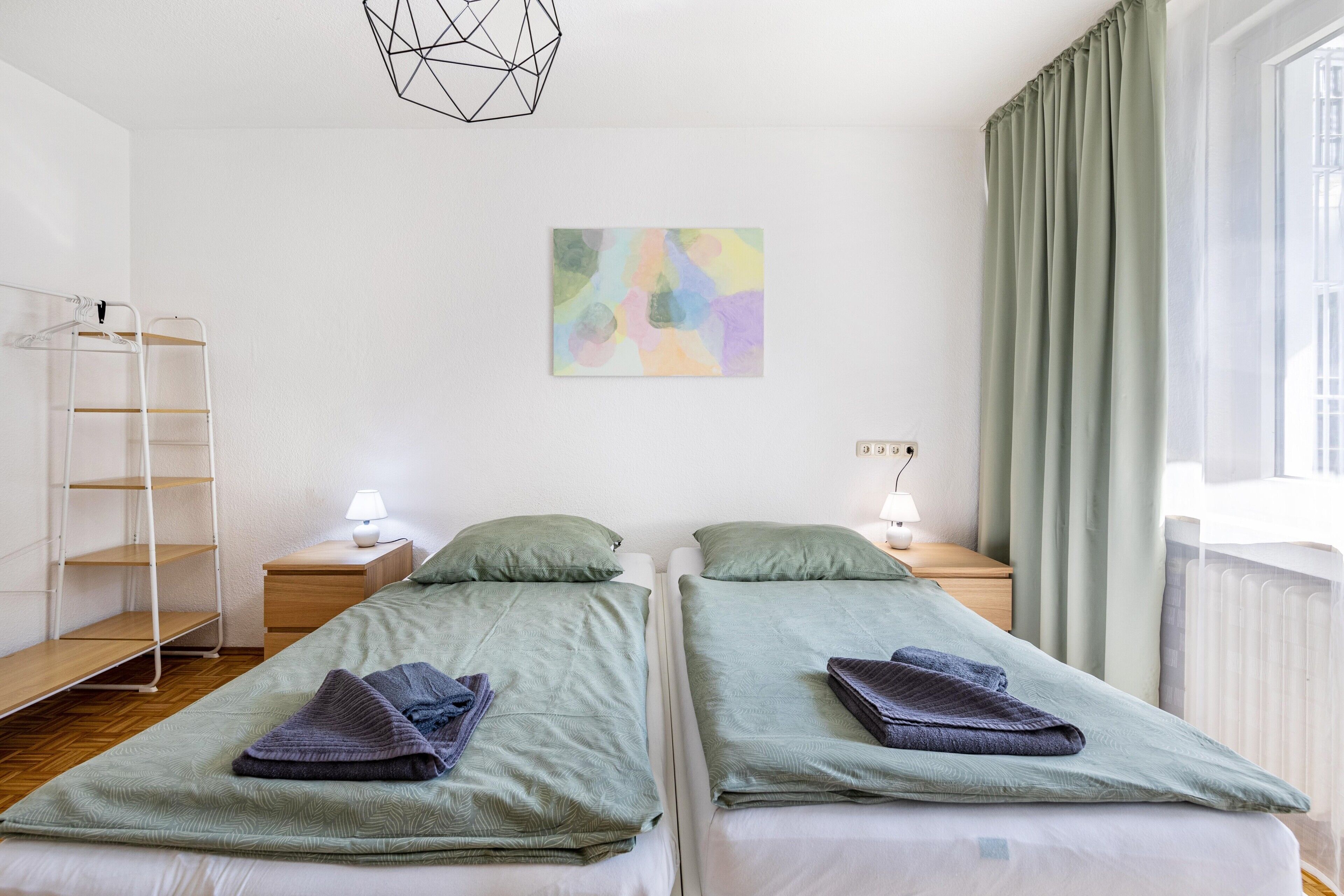 Ein Doppelzimmer mit zwei Betten, einem an der Wand befestigten abstrakten Gemälde, einem Fenster mit Vorhängen und einer geometrischen Leuchte.