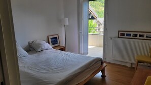 Um quarto com uma cama, uma mesa de cabeceira com abajur, um quadro emoldurado e vista para uma varanda com uma casa.