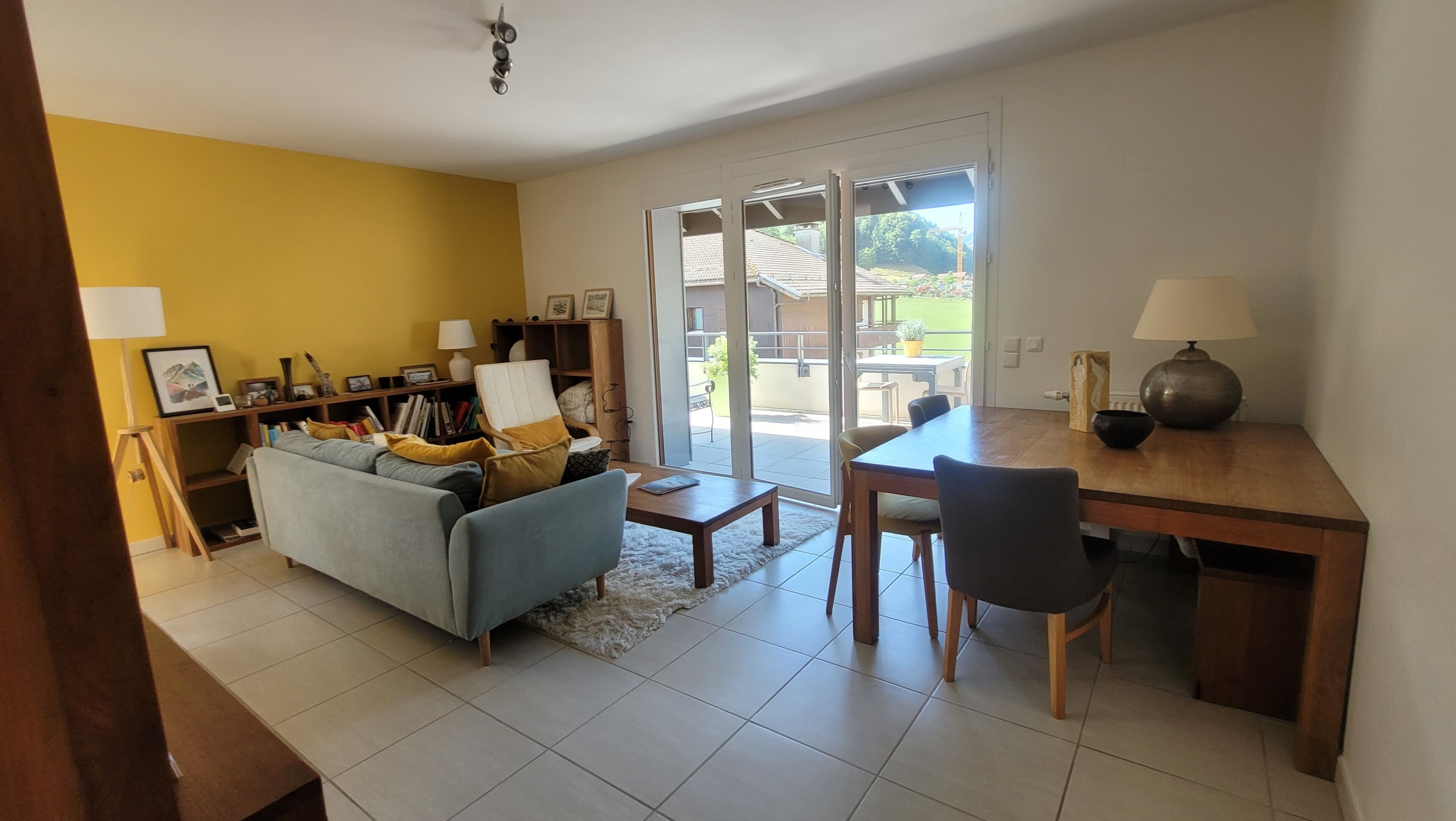 Una sala de estar con sofá, una mesa de centro y un comedor con mesa y sillas. Se observa una estantería y una lámpara.