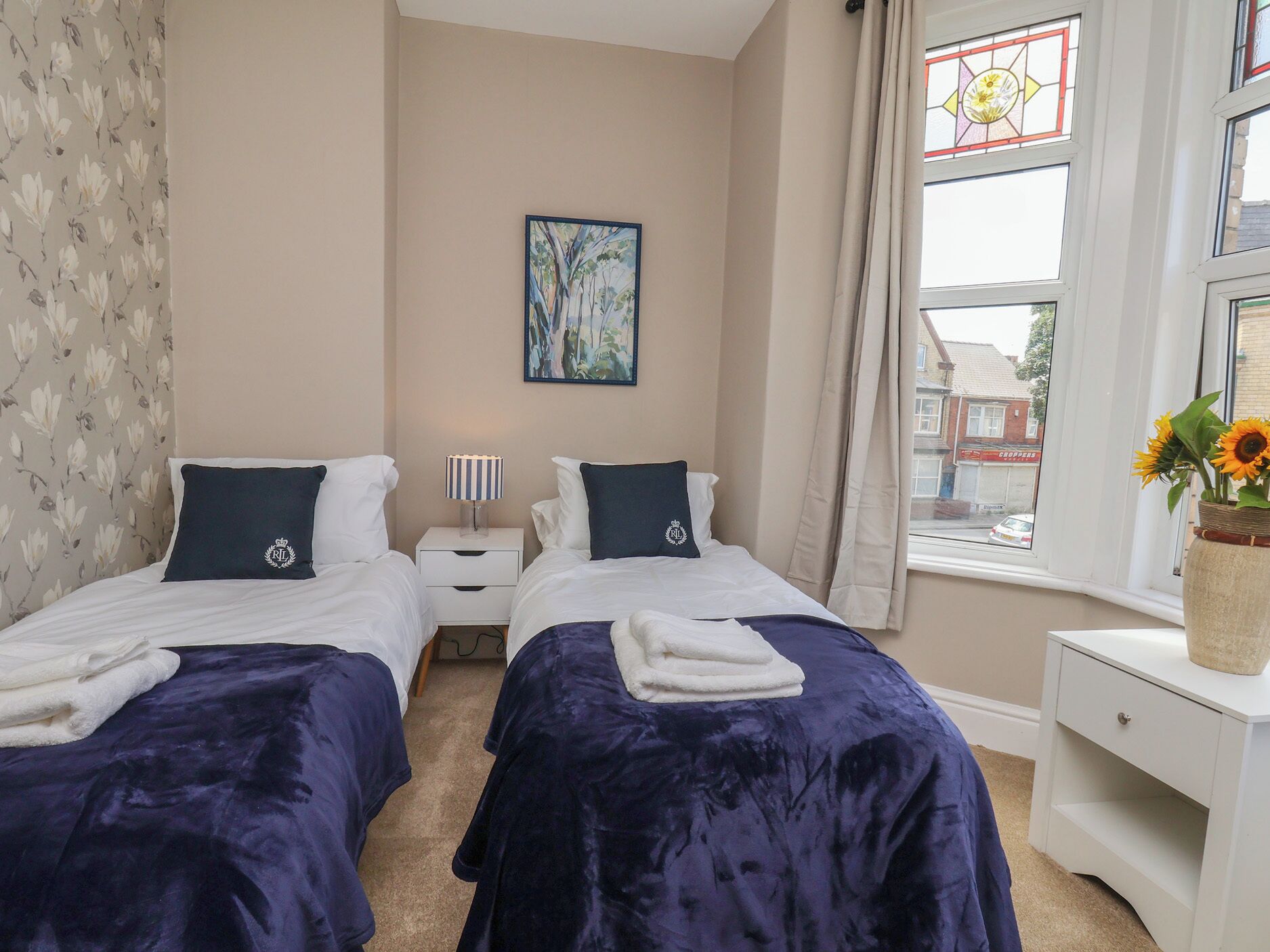 A bedroom with two beds, a nightstand with a lamp, a painting on the wall, and a window with a view of the outside.