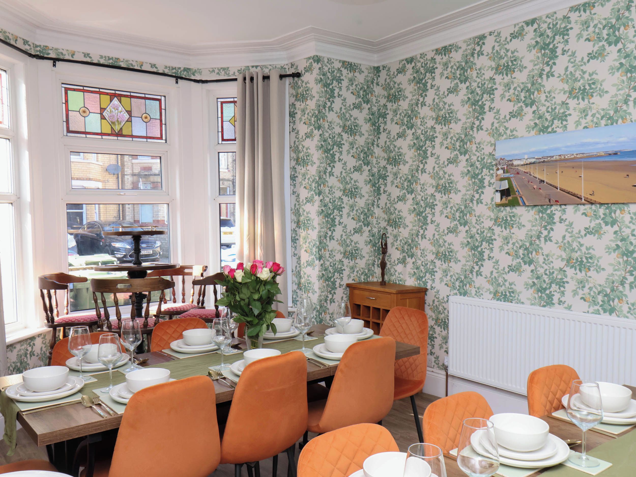 A dining area with a table set for four, surrounded by orange chairs, a vase of flowers, and a view of a beach through a window.