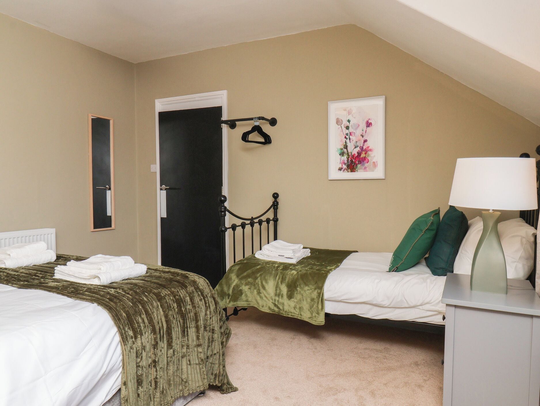 A bedroom with two beds, a door, a mirror, a framed artwork, and a bedside lamp.