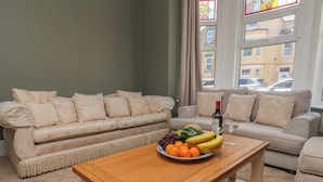 A living room with a beige sofa, a wooden coffee table with fruit and a bottle, and a view of buildings through a window.