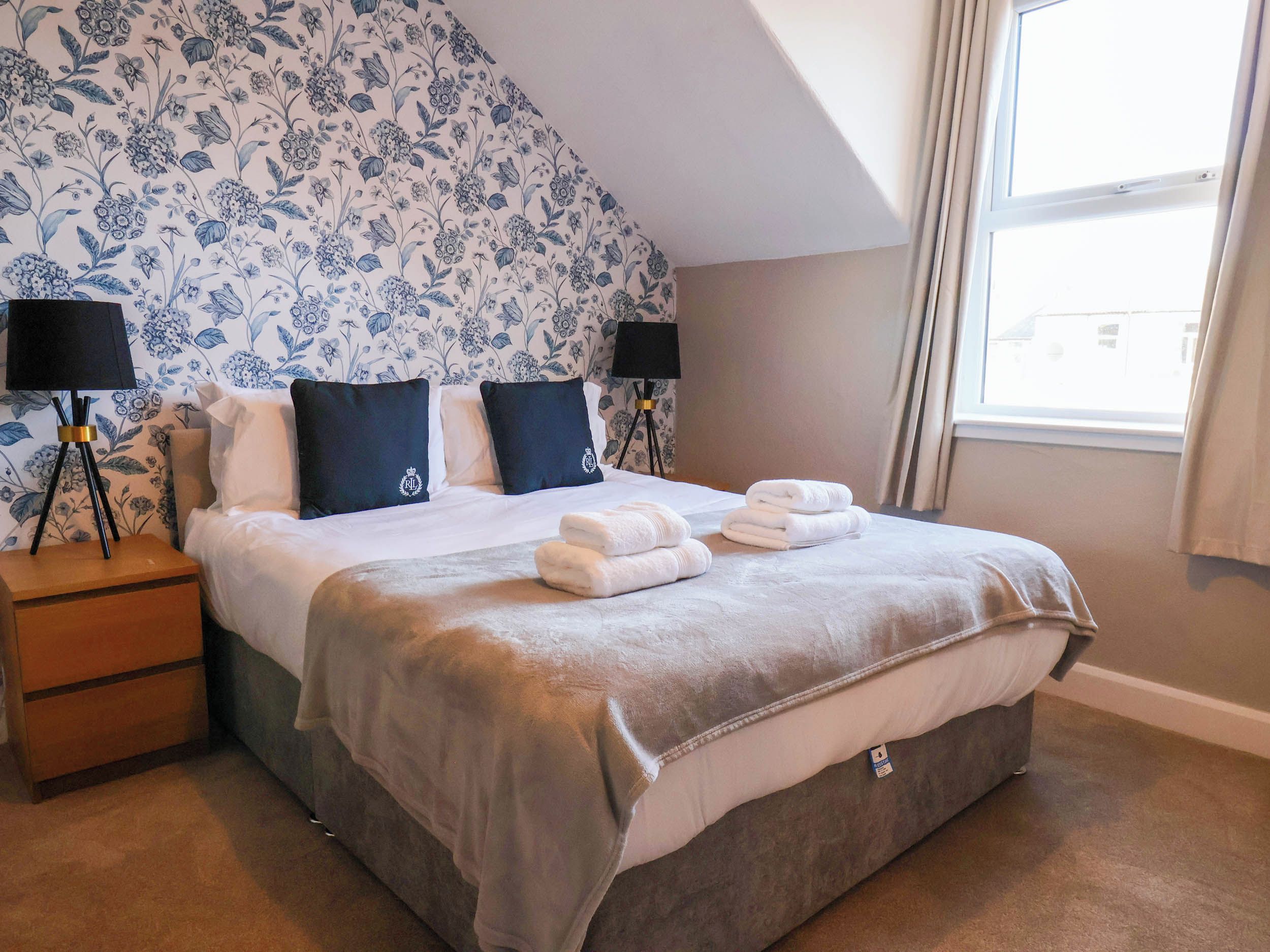 Habitación de hotel con cama, dos lámparas de noche, mesita auxiliar, ventana con cortinas y diseño floral en el papel tapiz.