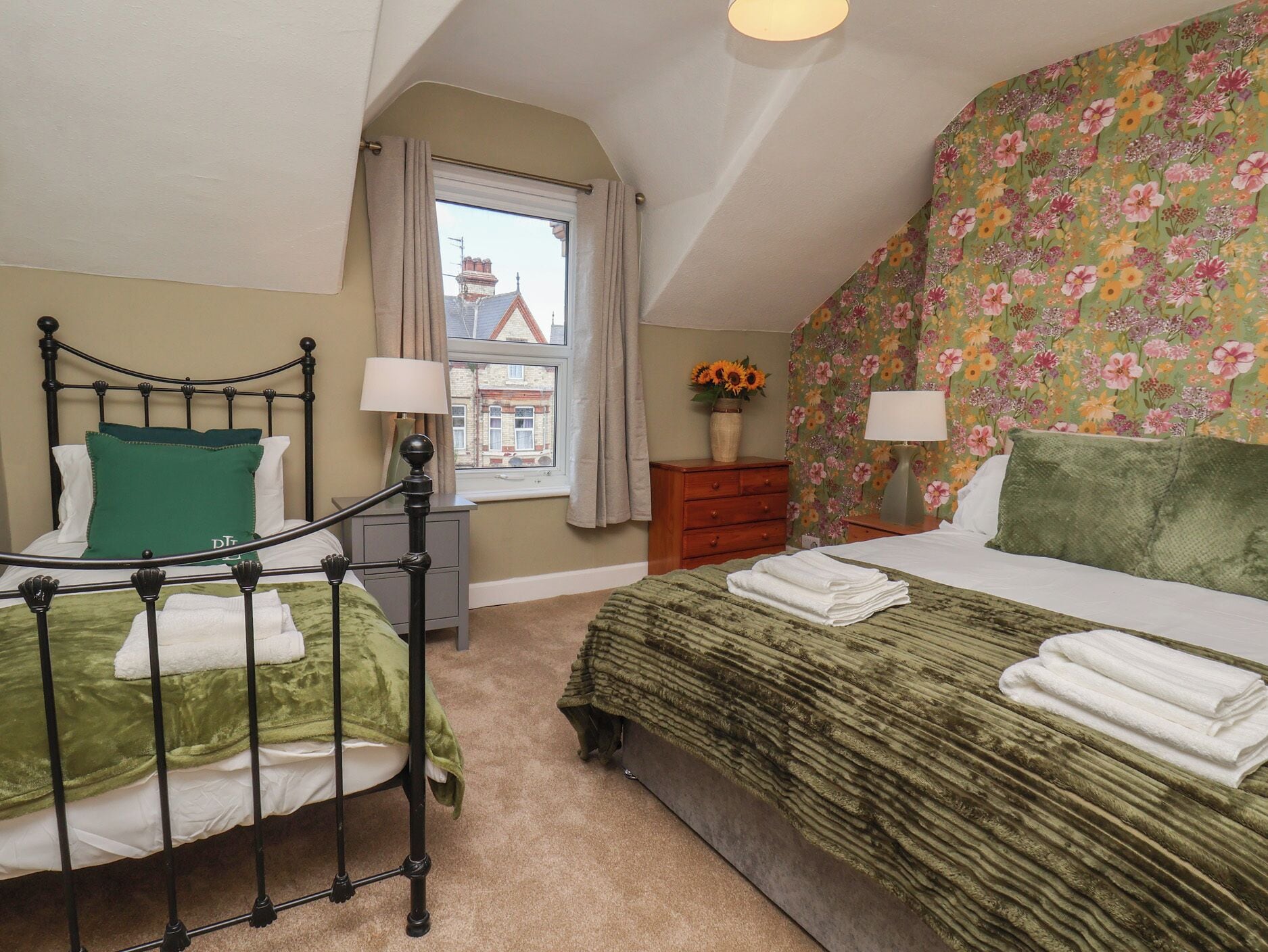 A bedroom with two beds, a window, a dresser, and a floral wallpaper.