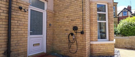 A corner of a brick building with a white door, a window, and a wall-mounted electrical box.