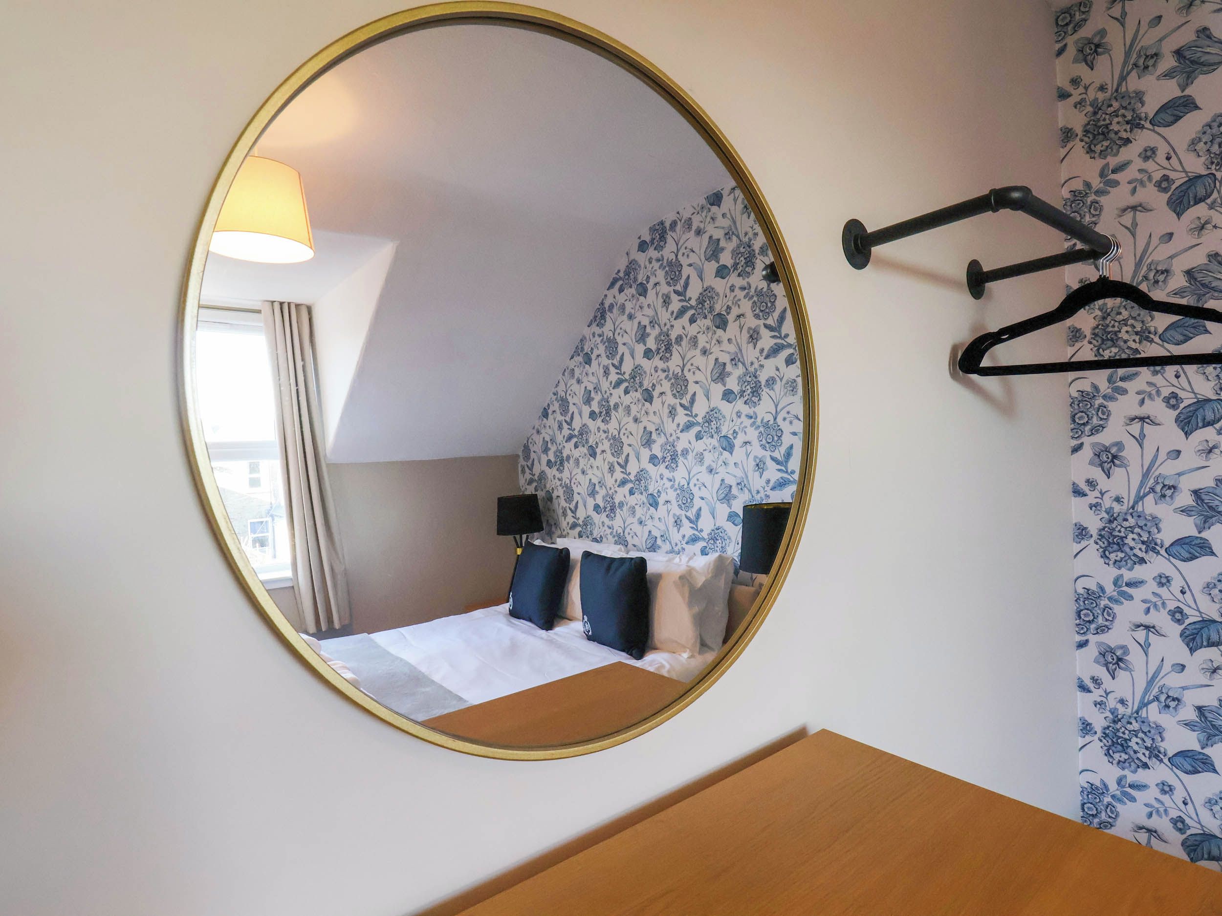 Une chambre avec un miroir rond, un lit avec une literie bleue et blanche, une table de chevet et un porte-manteau fixé au mur.