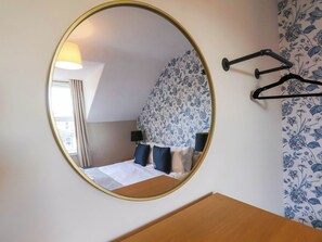 Une chambre avec un miroir rond, un lit avec une literie bleue et blanche, une table de chevet et un porte-manteau fixé au mur.
