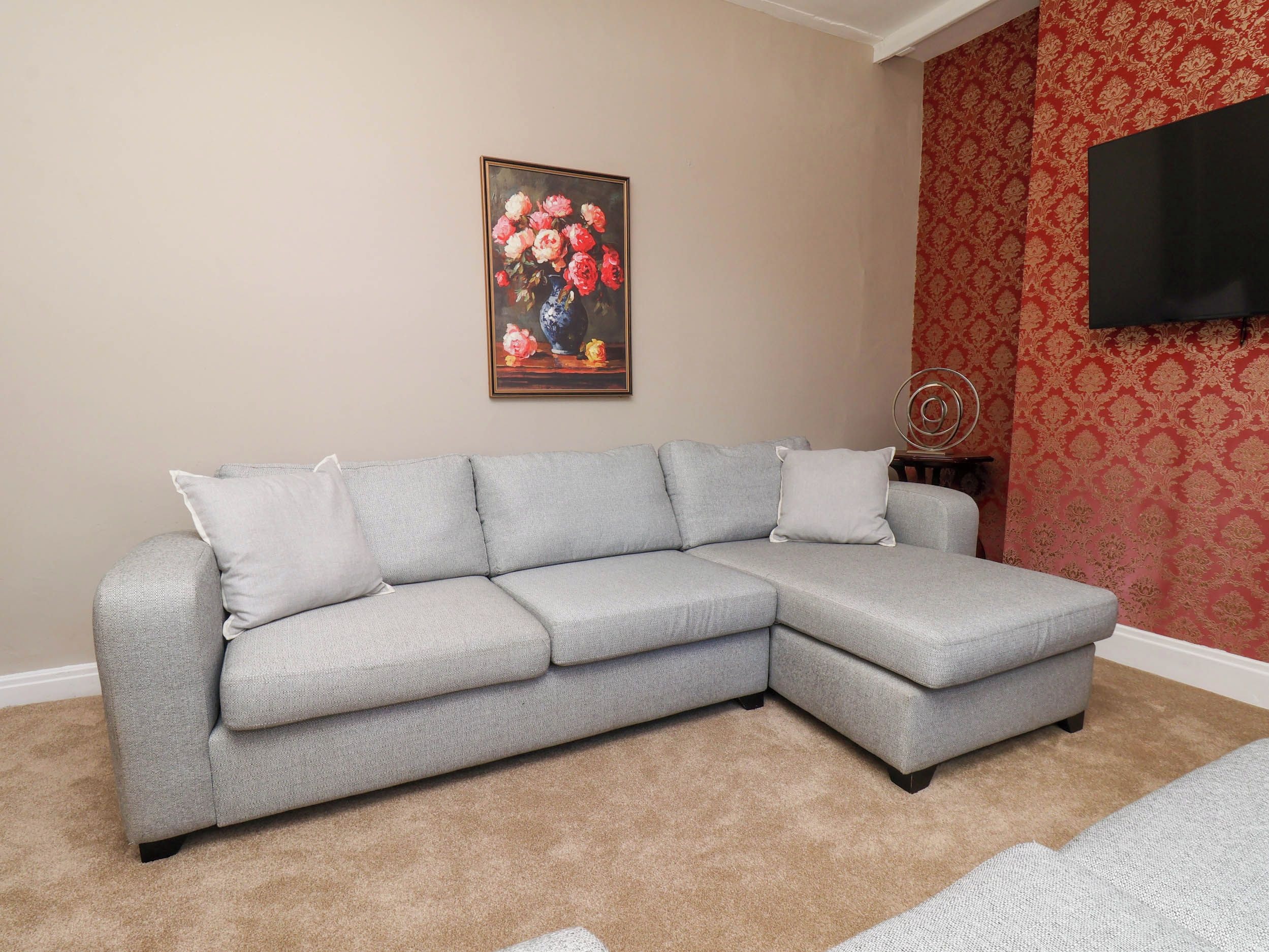 A grey sectional sofa with white cushions, a framed floral painting above it, and a red patterned wallpaper on the wall.