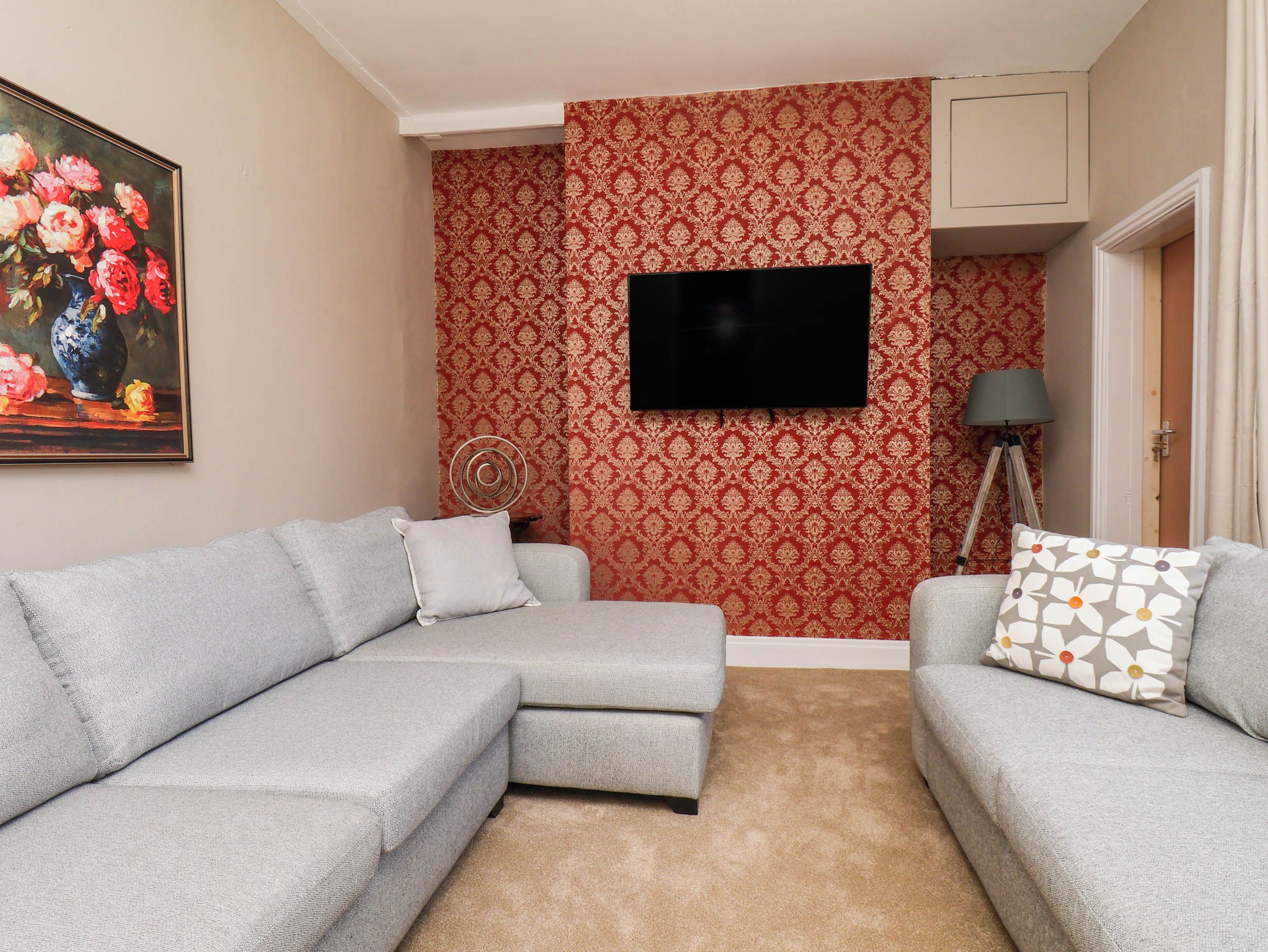 A living room with a sectional sofa, a flat-screen TV mounted on the wall, and patterned wallpaper.