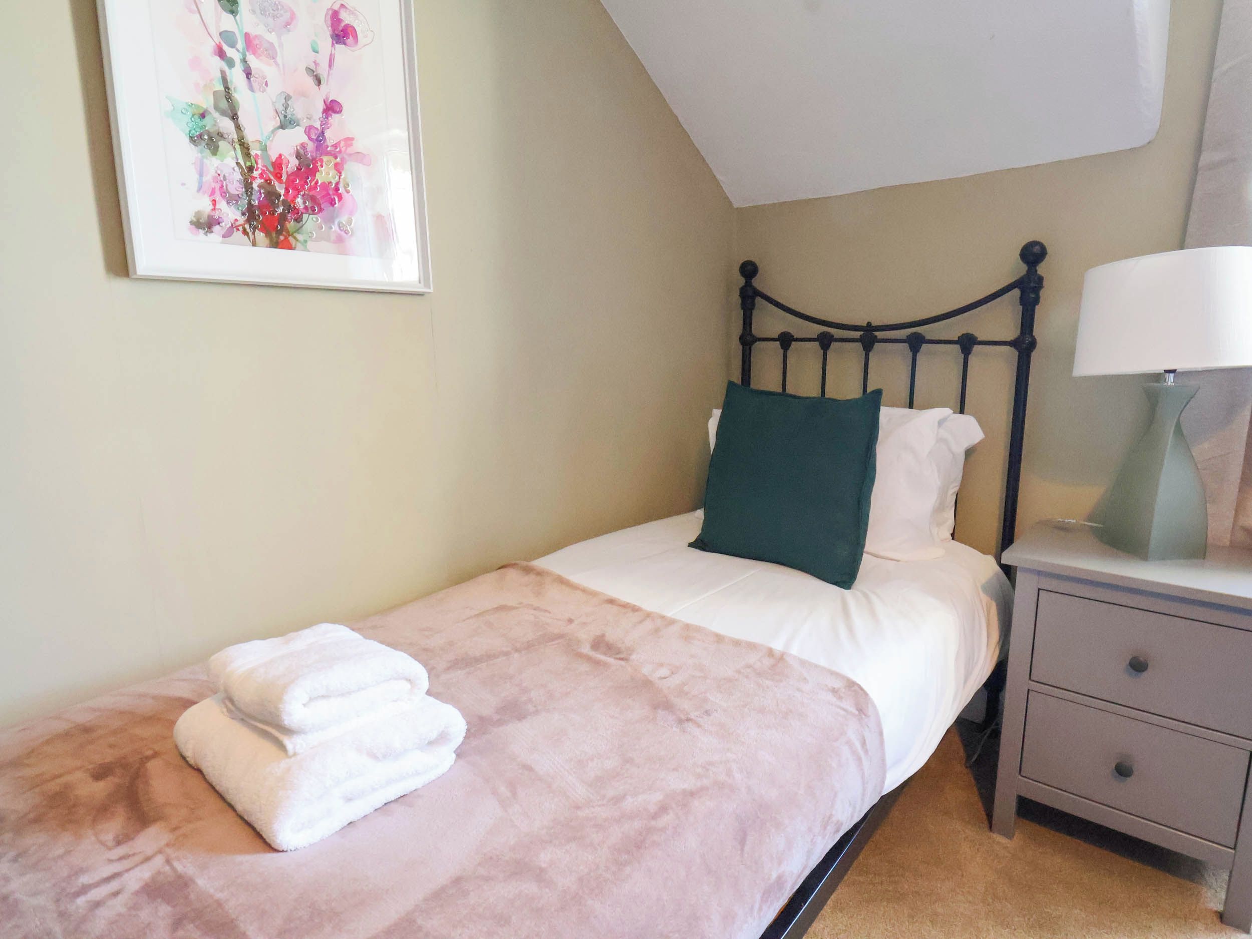 A single bed with a white and green pillow, a folded white towel, a bedside table with a lamp, and a framed floral artwork on the wall.
