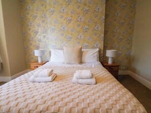 A neatly made bed with a patterned bedspread and two folded towels, flanked by bedside tables with lamps, in a room with floral wallpaper. - Lansdowne Grange (Bridlington)