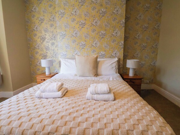Cama bem arrumada com colcha estampada e duas toalhas dobradas, com mesas de cabeceira com abajures, em um quarto com papel de parede floral.