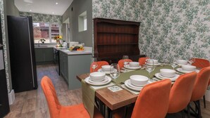 Ein Essbereich mit einem Holztisch, gedeckt für sechs Personen, umgeben von orangefarbenen Stühlen, einem dunklen Holzschrank und einer im Hintergrund sichtbaren Küche.