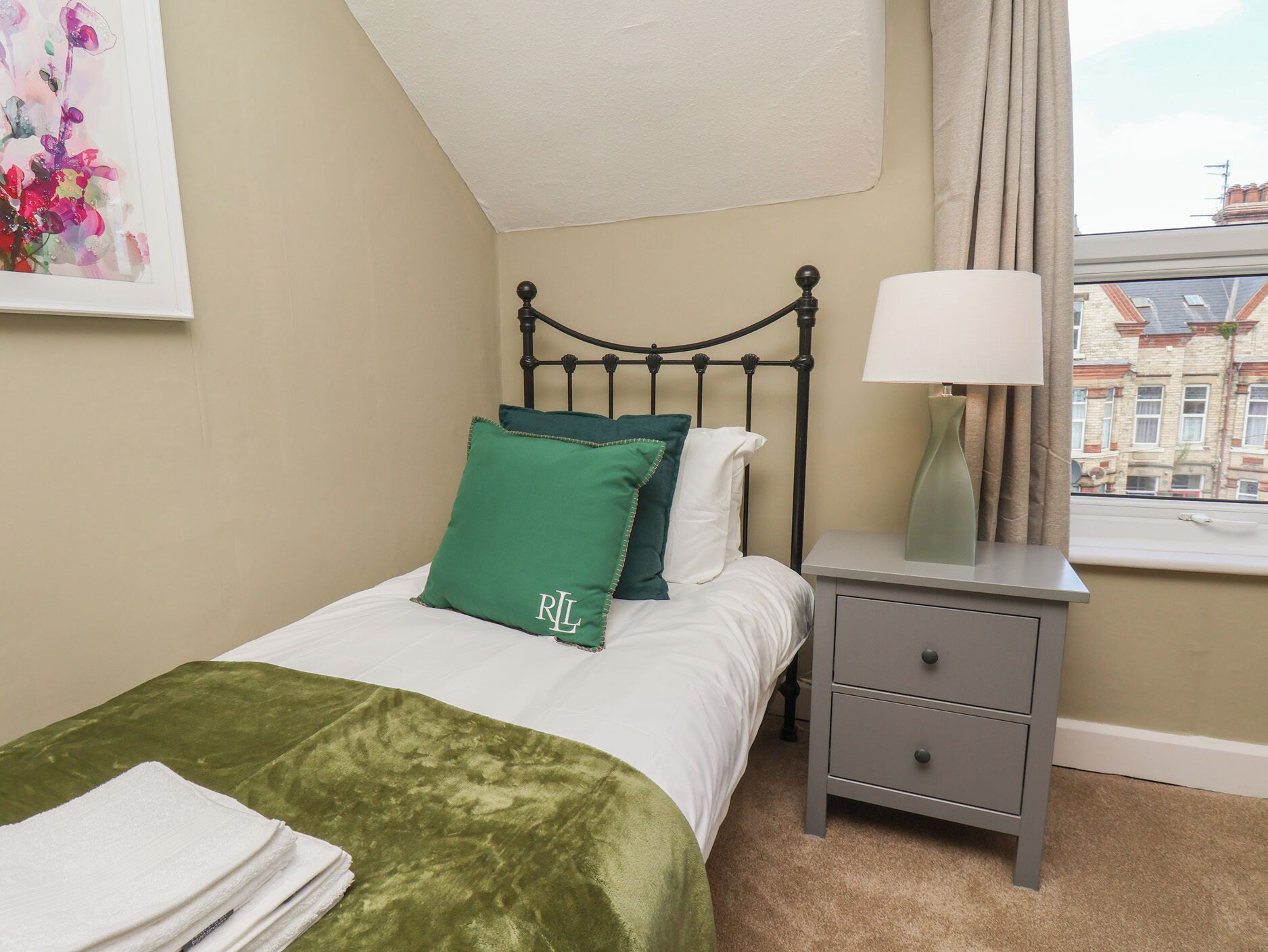 A bedroom with a bed, pillows, a bedside table with a lamp, a window with a view of buildings, and a painting on the wall.
