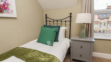 A bedroom with a bed, pillows, a bedside table with a lamp, a window with a view of buildings, and a painting on the wall.